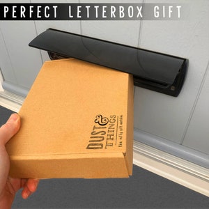 May include: A brown cardboard box with the text "Dust & Things" and "the nifty gift workshop" is being placed through a black letterbox. The text "PERFECT LETTERBOX GIFT" is at the top of the image. The letterbox is mounted on a white door.