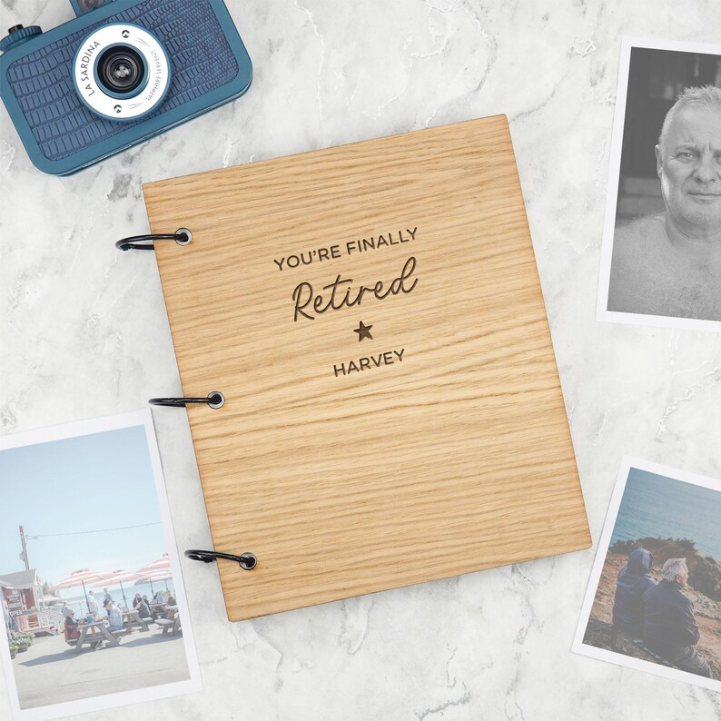 Puede incluir: Un &aacute;lbum de recortes rectangular de madera con tres anillas de metal negro en el lado izquierdo. La cubierta est&aacute; grabada con el texto "YOU'RE FINALLY Retired" seguido de un s&iacute;mbolo de estrella y el nombre "HARVEY" en letra oscura.