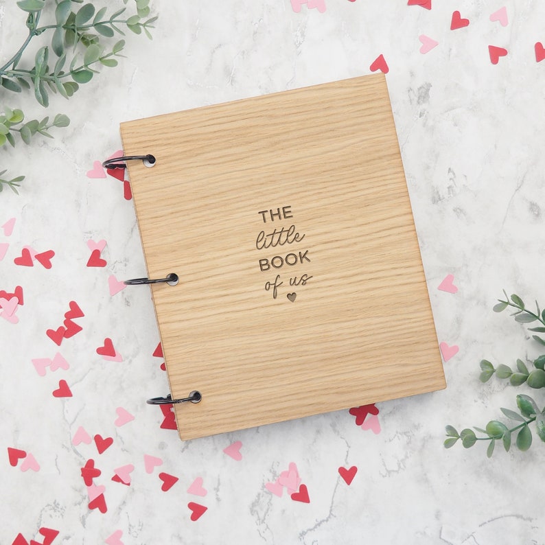 May include: A wooden journal with a ring binding. The cover is light brown and has the text "THE little BOOK of us" engraved on it. The text is in a cursive font and there is a small heart symbol below the text.
