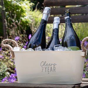 May include: A cream-colored metal beverage tub with two jute rope handles, filled with ice and three dark glass bottles of Brut sparkling wine. The front of the tub features the gray printed text Cheers to 1 Year with a small heart icon.