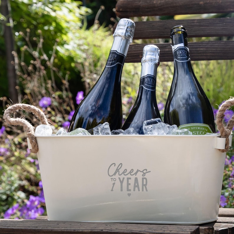 May include: A cream-colored metal beverage tub with two jute rope handles, filled with ice and three dark glass bottles of Brut sparkling wine. The front of the tub features the gray printed text Cheers to 1 Year with a small heart icon.