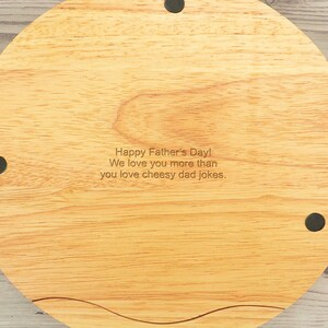 May include: A round wooden serving board with a natural wood grain finish. The board has a slightly curved edge and is engraved with the text "Happy Father's Day! We love you more than you love cheesy dad jokes."
