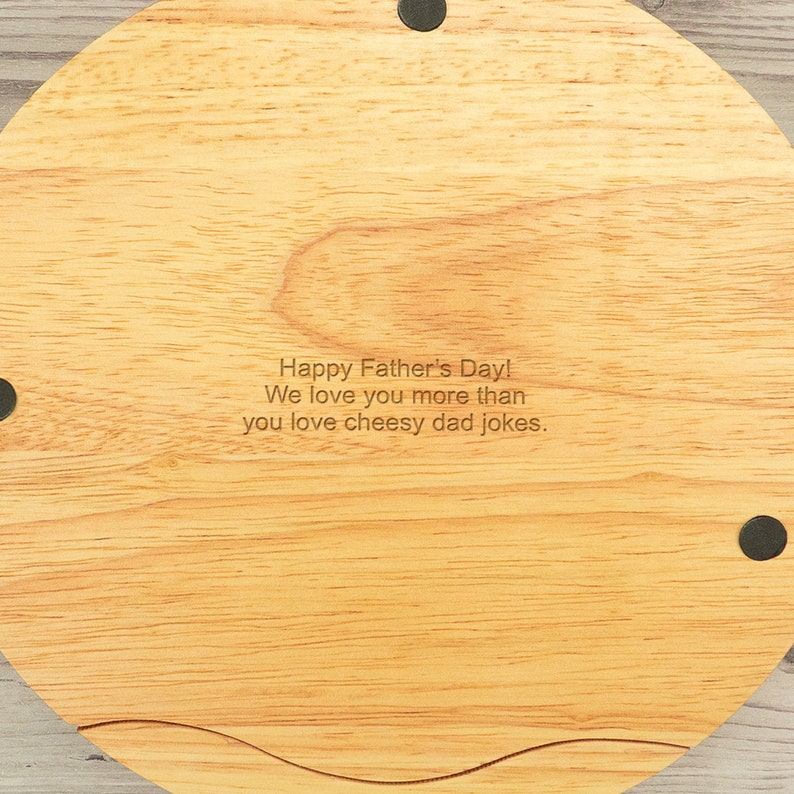 May include: A round wooden serving board with a natural wood grain finish. The board has a slightly curved edge and is engraved with the text "Happy Father's Day! We love you more than you love cheesy dad jokes."