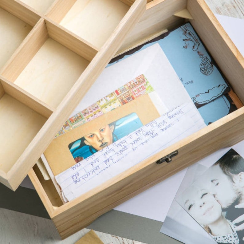 May include: A wooden box with a hinged lid, open to reveal a collection of letters, postcards, and photographs. The box is made of light wood and has a natural finish. The letters and postcards are handwritten and have a vintage feel. The photographs are black and white and show a couple.