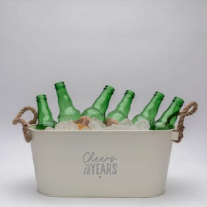 May include: An off-white oval metal beverage tub with two thick rope handles, filled with ice and six green glass bottles. The front of the tub features the gray printed text Cheers TO 18 YEARS with a small heart symbol centered below the text.