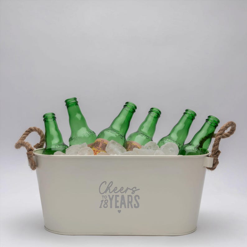 May include: An off-white oval metal beverage tub with two thick rope handles, filled with ice and six green glass bottles. The front of the tub features the gray printed text Cheers TO 18 YEARS with a small heart symbol centered below the text.