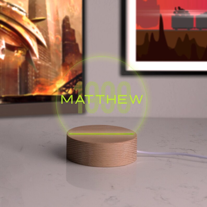 May include: A wooden LED night light with the name "MATTHEW" illuminated in green. The light sits on a round wooden base and is connected to a white cord. The background includes framed artwork.