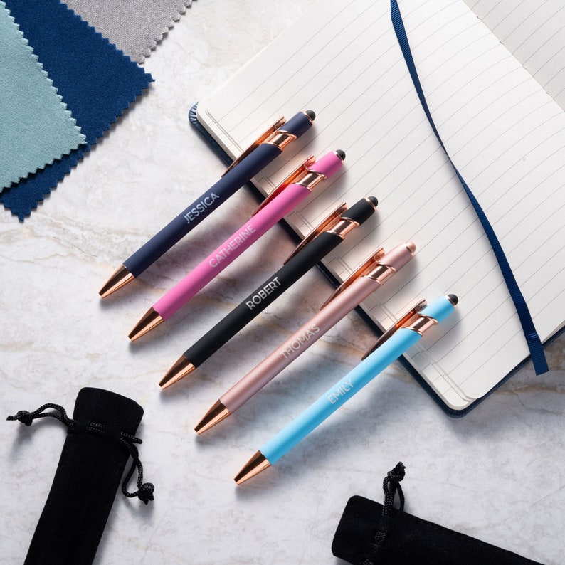 The image shows a collection of stylus pens laid out on a marble surface next to an open lined notebook. Each pen has a metallic finish, with what appears to be copper-colored tips and clips.