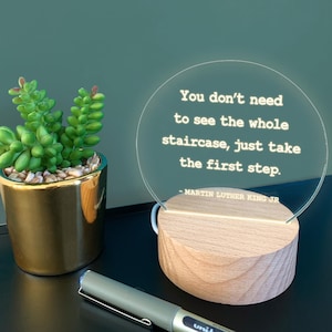 May include: An illuminated LED lamp with a wooden base and a clear, circular acrylic panel. The panel features the quote "You don't need to see the whole staircase, just take the first step." by Martin Luther King Jr. A succulent plant in a gold pot is visible.
