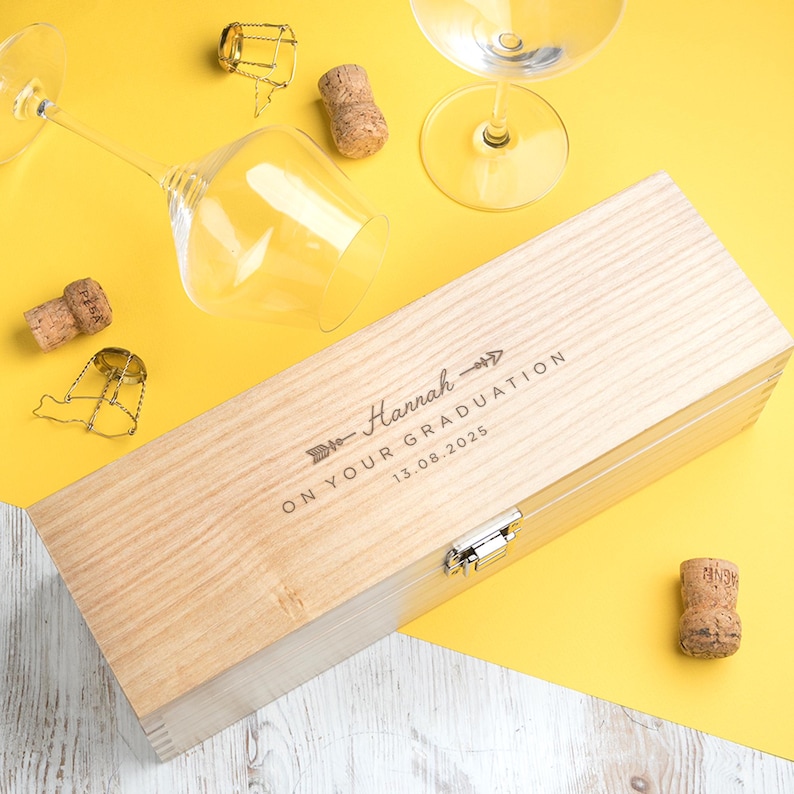 May include: A light-colored wooden wine box with the text "Hannah" and "ON YOUR GRADUATION 13.08.2025" engraved on the lid. The box is rectangular with a metal clasp. Wine glasses and corks are scattered around the box on a yellow surface.