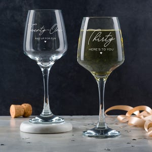 Puede incluir: Dos copas de vino de vidrio transparente con texto grabado. La copa de la izquierda dice "Twenty-One and Up For Fun" con un s&iacute;mbolo de coraz&oacute;n. La copa de la derecha dice "Thirty Here's To You" con un s&iacute;mbolo de coraz&oacute;n.