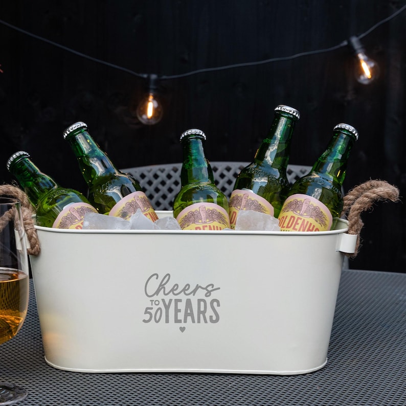 May include: An oval-shaped cream-colored metal beverage tub with two thick rope handles. The front features the gray text "Cheers to 50 YEARS" with a small heart icon. Inside, five green glass beer bottles are nestled in ice for cooling.