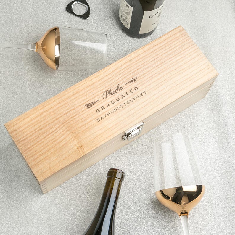 May include: A light-colored wooden wine box with the text "Phoebe GRADUATED BA (HONS) TEXTILES" is displayed with two wine glasses with gold rims, a bottle opener, and a bottle of wine.