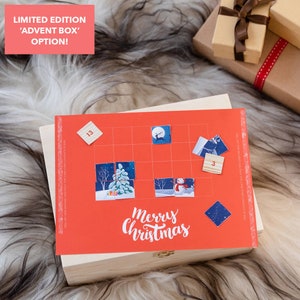 Puede incluir: Una caja de madera roja con un calendario de adviento de tem&aacute;tica navide&ntilde;a en la parte superior. El calendario tiene una cuadr&iacute;cula de cuadrados con n&uacute;meros e im&aacute;genes de un mu&ntilde;eco de nieve, un reno y un &aacute;rbol de Navidad. La caja tiene una tapa con bisagras y un peque&ntilde;o pestillo de metal. El texto "Feliz Navidad" est&aacute; impreso en el calendario.
