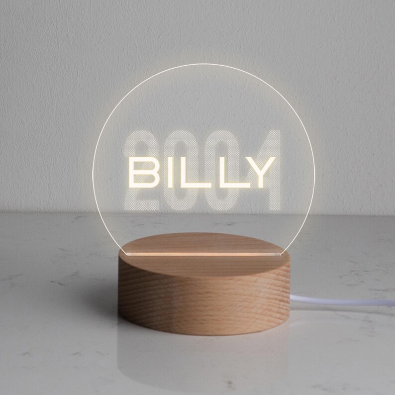May include: A personalized LED night light with the name "BILLY" illuminated in a soft, white glow. The design features a clear, circular acrylic panel set on a natural wood base. The year "2004" is faintly visible behind the name.