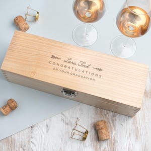 May include: A light-colored wooden wine box with the text "Lara Ford Congratulations On Your Graduation" engraved on the lid. Two champagne glasses and corks are visible. The box has a metal clasp.