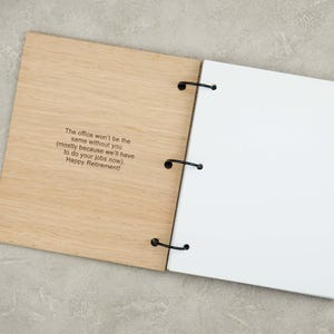 Puede incluir: Un &aacute;lbum de recortes de madera de color claro con tres anillas de metal negro que sujetan p&aacute;ginas blancas. La portada presenta el texto grabado: The office won't be the same without you (mostly because we'll have to do your jobs now). Happy Retirement!