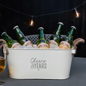 May include: An oval cream-colored metal beverage tub with two thick rope handles. The front features the gray text "Cheers TO 15 YEARS" with a small heart icon. Inside, five green glass beer bottles are nestled in ice to keep them chilled.