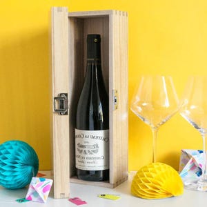 May include: A bottle of wine in a wooden gift box with a silver clasp. The wine bottle has a white label with black text. Two wine glasses and decorative paper balls are in the background. The background is yellow.