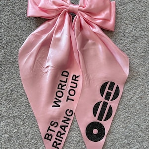Pode incluir: Um grande laço de cetim rosa claro com caudas longas e pontiagudas. As caudas apresentam o texto "BTS WORLD ARIRANG TOUR" em preto, juntamente com um gráfico preto. Provavelmente um acessório de cabelo ou um item decorativo.