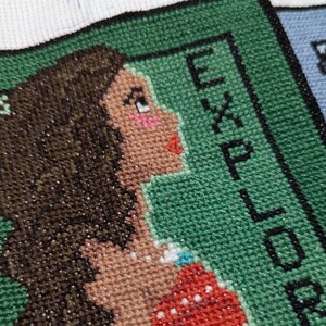 Puede incluir: Un bordado de punto de cruz verde con un retrato de una mujer con cabello castaño y una blusa roja. El texto "EXPLORER" es visible en el lado derecho de la imagen.