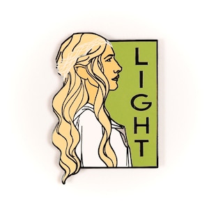 Può includere: Uno spillo in smalto verde e nero con un disegno a linea di una donna con lunghi capelli biondi e la parola "LIGHT" verticalmente sul lato destro.