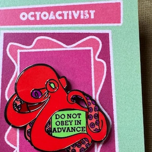 May include: A red octopus enamel pin with the text "DO NOT OBEY IN ADVANCE" on a green background. The pin is on a pink and green card with the text "OCTOACTIVIST" at the top and artist information at the bottom.