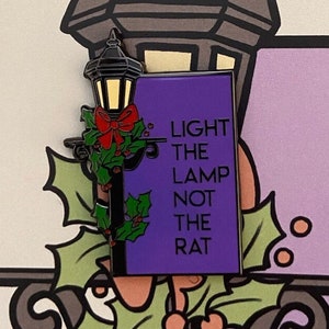 May include: A purple enamel pin with a black outline featuring a street lamp with a red bow and holly. The text on the pin reads "Light the lamp not the rat".
