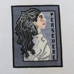 Può includere: Ricamo a punto croce di una donna con lunghi capelli scuri e un disegno floreale nei capelli. Lo sfondo è grigio chiaro con la parola "PERSEVERE" scritta verticalmente in nero.