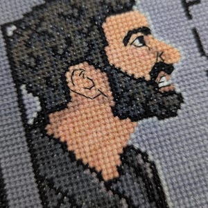 Peut inclure: Portrait en point de croix d'un homme avec une barbe et des cheveux foncés, rendu en nuances de noir, de gris et de beige. L'image est un gros plan du visage de l'homme de profil, avec la lettre "F" partiellement visible en arrière-plan.