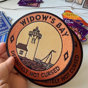 Puede incluir: Un parche bordado redondo que muestra un faro y un velero en una costa rocosa. El borde es naranja con texto negro que dice WIDOW'S BAY y DEFINITELY NOT CURSED. El centro es un círculo amarillo claro con una ilustración de un faro negro.
