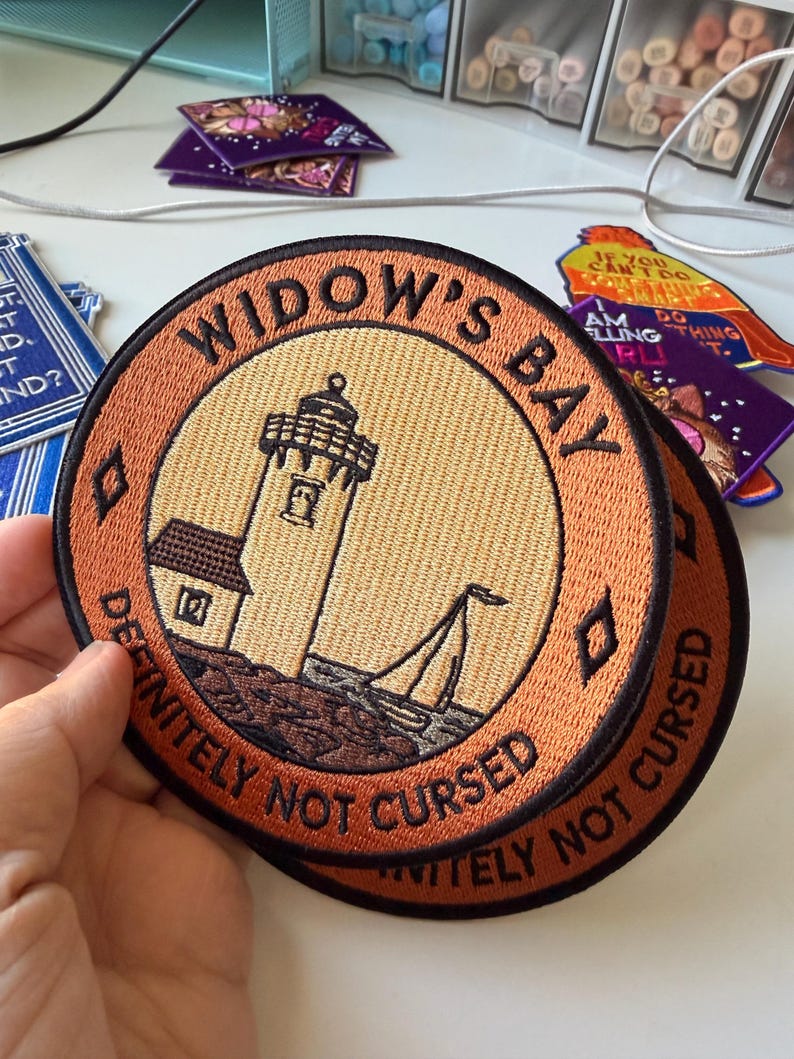 Op de afbeelding: Een ronde geborduurde patch met een vuurtoren en een zeilboot op een rotsachtige kust. De oranje rand bevat zwarte tekst met WIDOW'S BAY en DEFINITELY NOT CURSED. Het midden is een lichtgele cirkel met een illustratie van een vuurtoren.