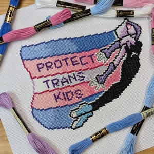 Może przedstawiać: Haft krzyżykowy z napisem "PROTECT TRANS KIDS" w kolorze różowym i białym, na niebiesko-różowym tle. Projekt zawiera stylizowaną postać z wyciągniętymi ramionami. Wokół pracy nici do haftu w różnych kolorach.
