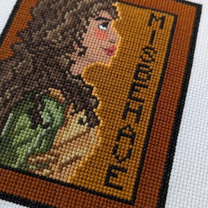 May include: Cross-stitch embroidery of a woman with brown hair in a green and brown dress, framed in brown and black. The text "MISSISSIPPI" is stitched in black on the right side of the frame.