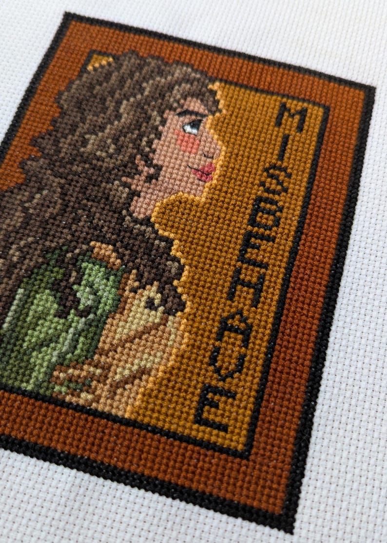 May include: Cross-stitch embroidery of a woman with brown hair in a green and brown dress, framed in brown and black. The text "MISSISSIPPI" is stitched in black on the right side of the frame.