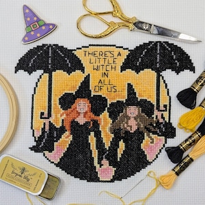 Può includere: Un disegno a punto croce con due streghe sotto ombrelli, con la scritta "THERE'S A LITTLE WITCH IN ALL OF US...". Il disegno è circondato da forniture per ricamo, tra cui forbici, filo e una piccola scatola di balsamo per filo di cera d'api.