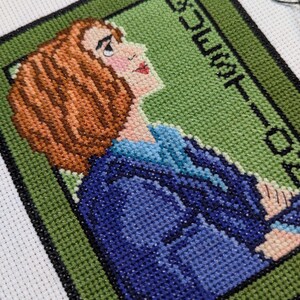 May include: Cross-stitch embroidery of a woman with brown hair wearing a blue jacket. The background is green with the text "COUNT-DOWN" in black.