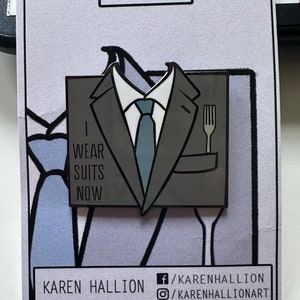May include: A gray enamel pin featuring a cartoon suit with a tie and a fork in the pocket. The text "I wear suits now" is printed on the suit. The pin is on a white card with the text "FORKS" at the top and "KAREN HALLION" and social media handles at the bottom.