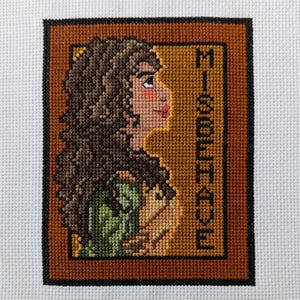 May include: Cross-stitch embroidery of a woman with brown hair, wearing a green dress, with the word "Misbehave" stitched in brown thread on a gold background.