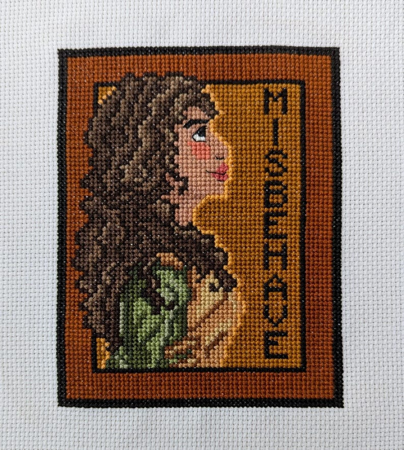 May include: Cross-stitch embroidery of a woman with brown hair, wearing a green dress, with the word "Misbehave" stitched in brown thread on a gold background.