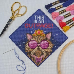 May include: A cross-stitch project on white fabric featuring a cat wearing a gold crown and pink sunglasses, with the text THIS IS AN OUTRAGE in red and white. Includes gold-tone scissors, a needle with blue thread, and colourful embroidery floss.