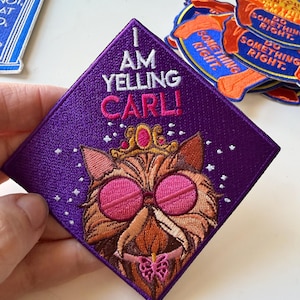 May include: A diamond-shaped purple embroidered patch featuring a cat wearing a gold crown and pink circular glasses. The text reads I AM YELLING CARL! in white and pink thread. Small white stars surround the cat, and a pink butterfly is at the bottom.