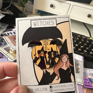 May include: A pin featuring three witches under a black umbrella, with the text "WITCHES" at the top. The pin has the text "THERE'S A LITTLE WITCH IN ALL OF US..." and the artist's social media handles.