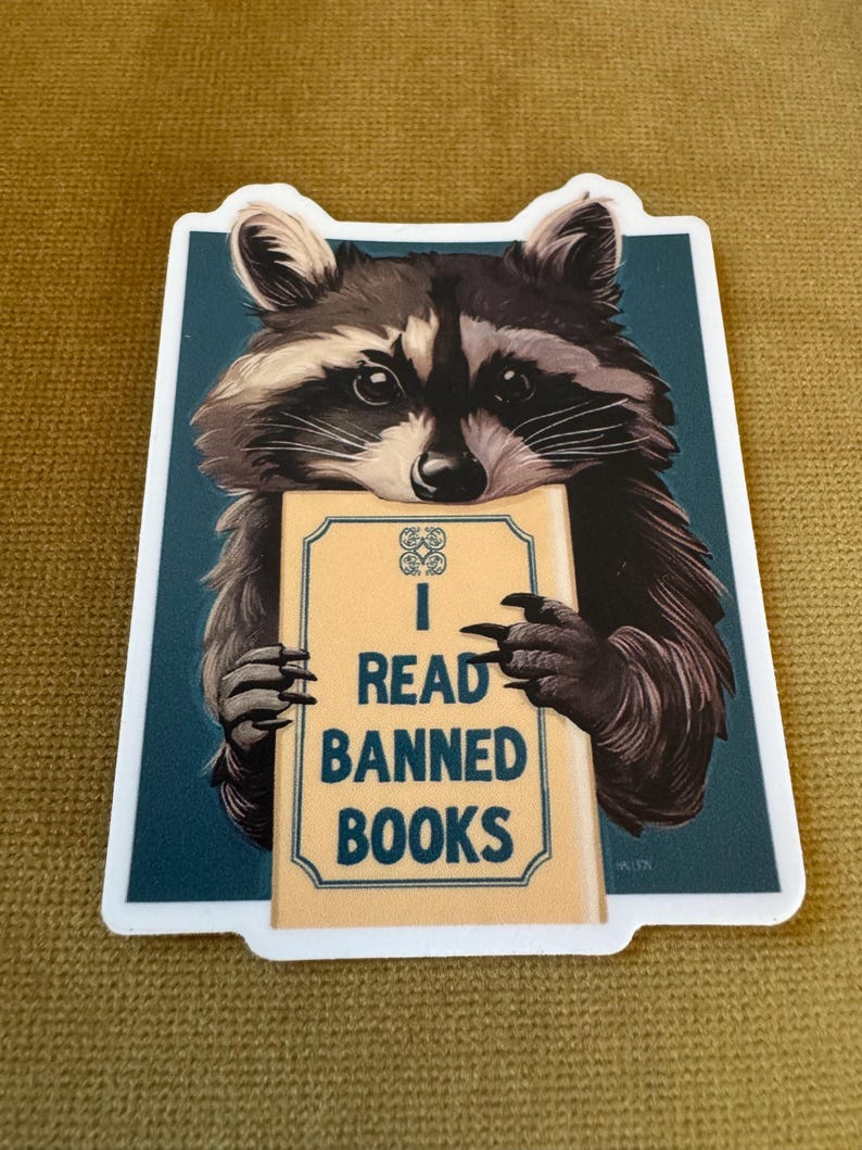 May include: A rectangular sticker featuring a detailed illustration of a raccoon with black and grey fur holding a tan book. The book cover displays a decorative emblem and the text I READ BANNED BOOKS in dark blue capital letters.