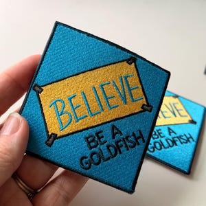 May include: A diamond-shaped embroidered patch with a bright blue background and a central yellow rectangular banner. The text BELIEVE is stitched in blue on the yellow banner, and the words BE A GOLDFISH are stitched in black below it.