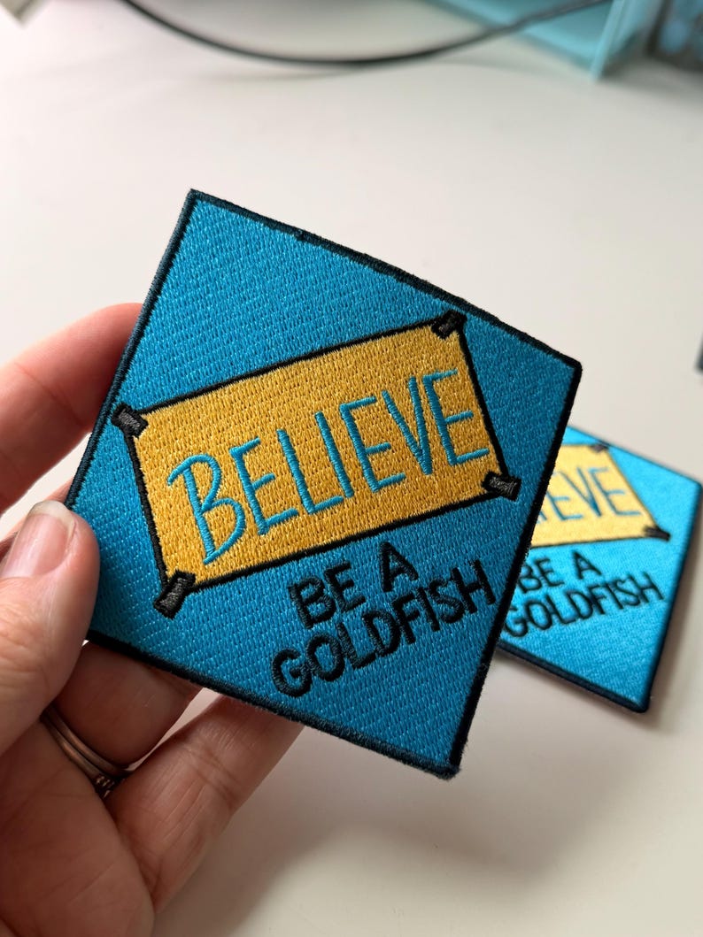 May include: A diamond-shaped embroidered patch with a bright blue background and a central yellow rectangular banner. The text BELIEVE is stitched in blue on the yellow banner, and the words BE A GOLDFISH are stitched in black below it.