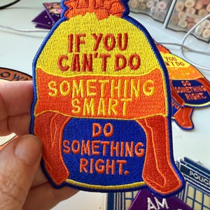 Peut inclure: Un écusson brodé en forme de bonnet d'hiver avec une bordure bleue. Le bonnet est jaune et orange avec le texte IF YOU CAN'T DO SOMETHING SMART DO SOMETHING RIGHT. Le motif présente une section de base bleue et un pompon orange au sommet.