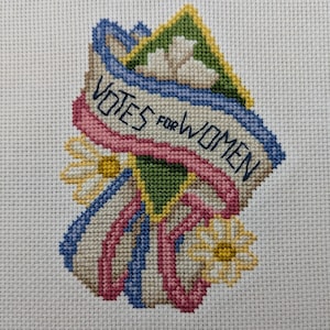 May include: Cross-stitch design with the text "VOTES FOR WOMEN" on a banner, surrounded by daisies and ribbons in blue, pink, and green. The needlework is on a white fabric background.