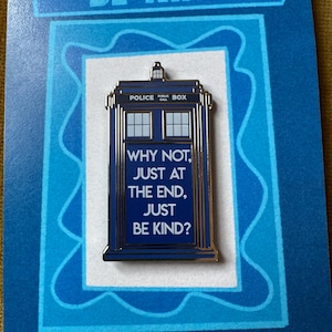 Op de afbeelding: Een blauwe emaille pin in de vorm van een politiebox met de tekst "WHY NOT, JUST AT THE END, JUST BE KIND?". De pin zit op een blauwe kaart met bovenaan de tekst "BE KIND".