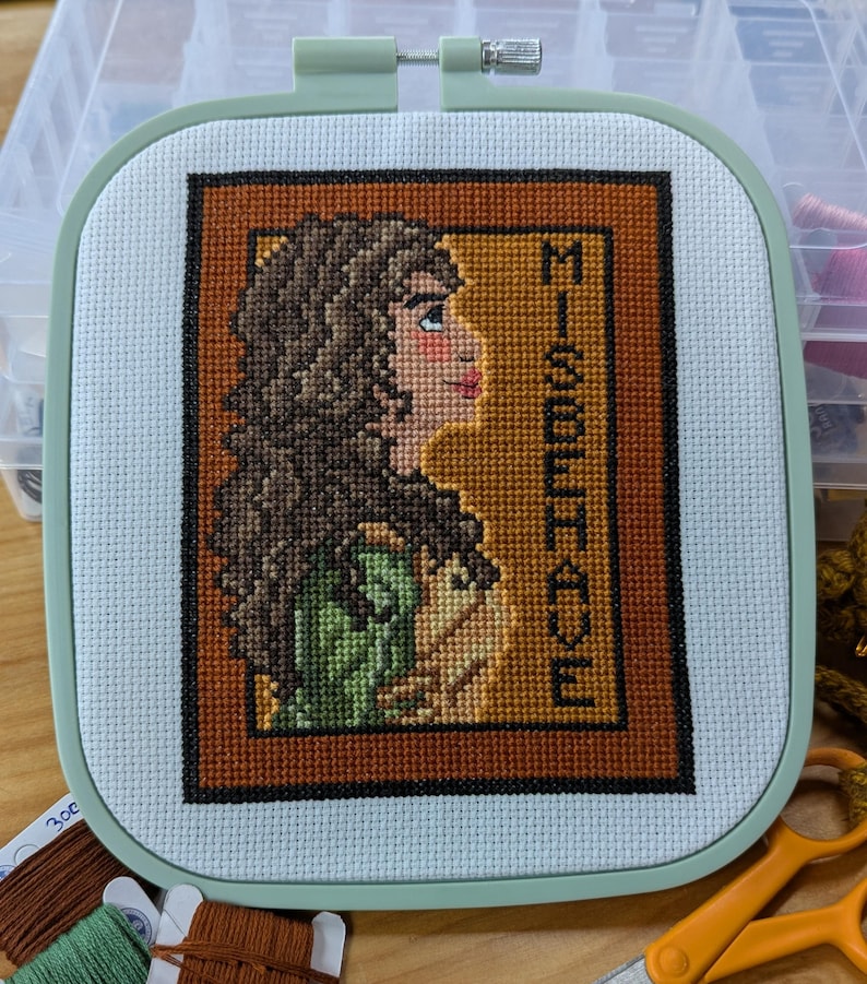 May include: A cross-stitch embroidery hoop with a framed image of a woman with brown hair and a green dress. The text "MISBEHAVE" is stitched in the background.